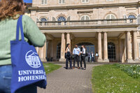 Foto: Eine Person mit einer blauen Tasche mit dem Logo der Universität Hohenheim steht vor Schloss Hohenheim. Mehrere Personen unterhalten sich vor dem Gebäude.