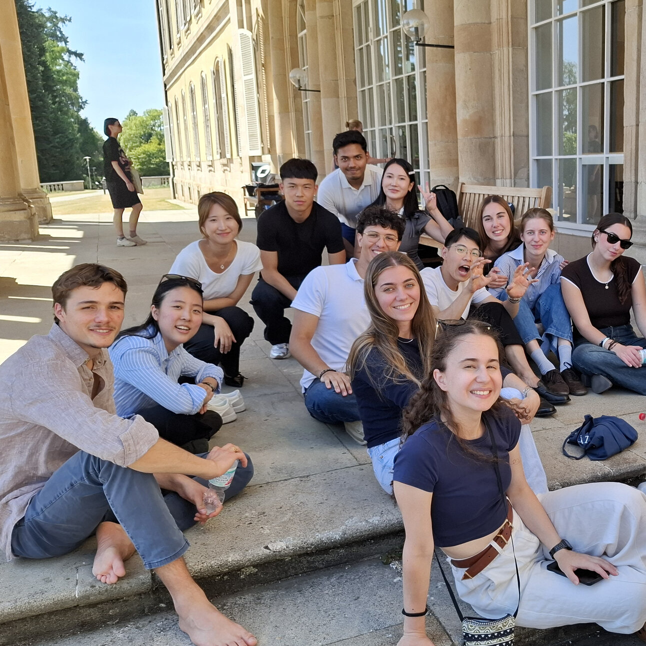 Foto: Eine Gruppe von Personen sitzt auf den Stufen vor einem großen Gebäude mit Säulen und Fenstern. Einige Personen halten Wasserflaschen. Die Umgebung wirkt sonnig und freundlich.
