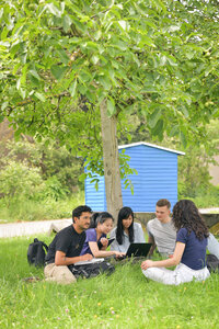 Foto: Eine Gruppe von Personen sitzt auf einer Wiese unter einem Baum. Sie sind um einen Laptop versammelt und scheinen zu arbeiten oder zu lernen. Im Hintergrund ist ein blaues Gebäude sichtbar.
