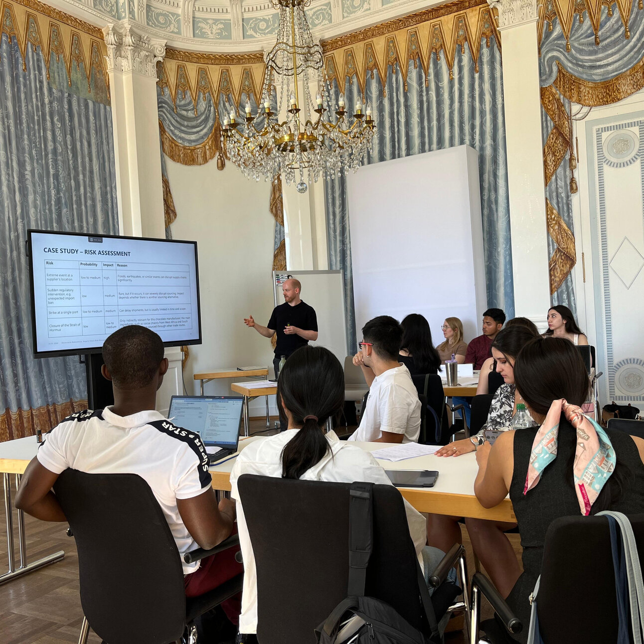 Foto: Eine Gruppe von Personen sitzt in einem dekorativen Raum und hört einem Vortrag über Risikobewertung zu. Auf einem Bildschirm ist der Titel 'CASE STUDY - RISK ASSESSMENT' zu sehen.
