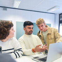 Foto: Eine Gruppe von Personen sitzt an einem Tisch in einem Büro. Eine Person zeigt auf einen Laptop, während andere Notizen machen. Bücher und Dokumente liegen auf dem Tisch.
