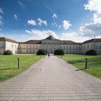 Foto: Schloss Hohenheim mit symmetrischem Design und Kuppeldach, umgeben von gepflegtem Rasen unter blauem Himmel mit Wolken. Zwei Personen gehen auf dem Weg zum Eingang.