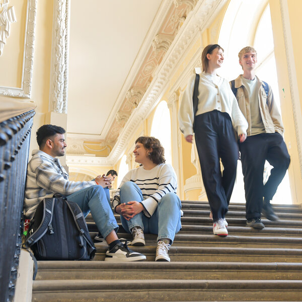 Foto: Mehrere Personen sitzen und stehen auf einer breiten Treppe in einem Gebäude mit dekorativen Wänden und hohen Decken. Zwei Personen sitzen auf den Stufen, während zwei andere die Treppe hinuntergehen.