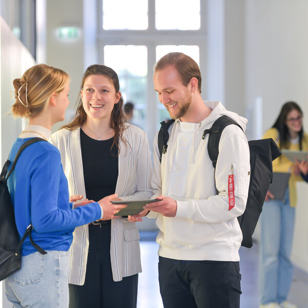 Foto: Drei Personen stehen in einem Flur und unterhalten sich. Eine Person hält ein Tablet. Im Hintergrund sind zwei weitere Personen zu sehen, die ebenfalls ein Tablet betrachten.