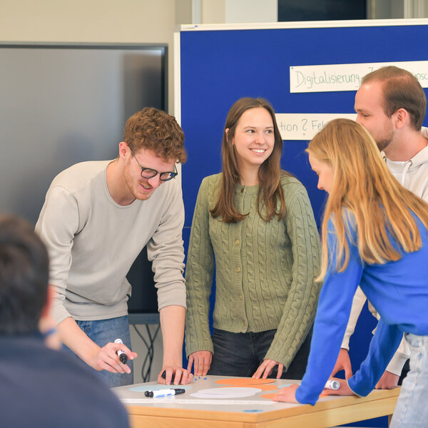 Foto: Vier Personen stehen um einen Tisch und arbeiten zusammen. Im Hintergrund ist eine Tafel mit dem Text 'Digitalisierung' sichtbar. Eine weitere Person sitzt im Vordergrund.