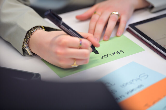 Foto: Eine Person schreibt mit einem schwarzen Stift auf eine grüne Karteikarte. Der Text auf der Karte lautet 'Honour'. Auf dem Tisch liegen weitere bunte Karteikarten und ein Tablet.