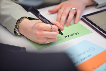Foto: Eine Person schreibt mit einem schwarzen Stift auf eine grüne Karteikarte. Der Text auf der Karte lautet 'Honour'. Auf dem Tisch liegen weitere bunte Karteikarten und ein Tablet.