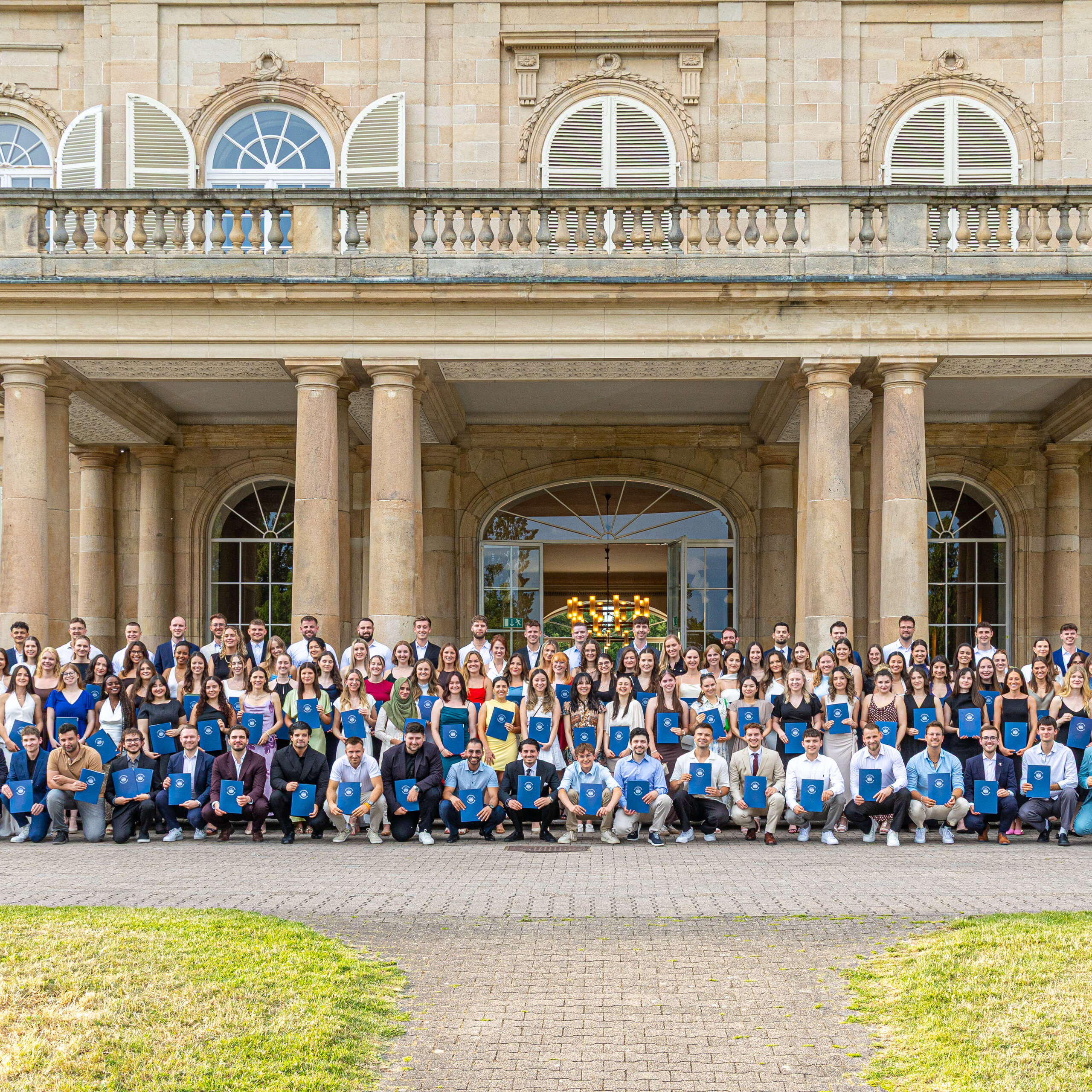 Foto: Eine große Gruppe von Personen steht vor Schloss Hohenheim. Die Personen halten blaue Mappen und posieren für ein Gruppenfoto vor dem historischen Gebäude mit Säulen und symmetrischem Design.