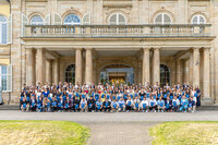 Foto: Eine große Gruppe von Personen steht vor Schloss Hohenheim. Die Personen halten blaue Mappen und posieren für ein Gruppenfoto vor dem historischen Gebäude mit Säulen und symmetrischem Design.