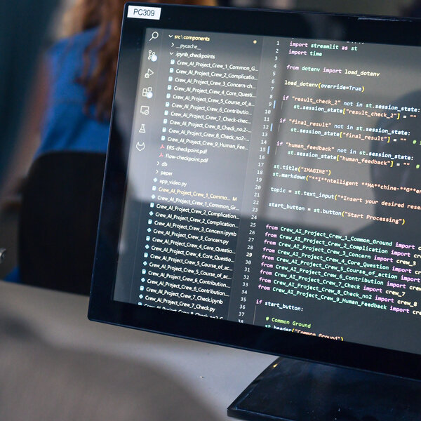 Foto: Ein Computerbildschirm zeigt Code in einer Entwicklungsumgebung. Im Hintergrund ist eine Person unscharf zu sehen. Der Code enthält Importanweisungen und Variablen für ein Projekt.