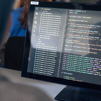 Foto: Ein Computerbildschirm zeigt Code in einer Entwicklungsumgebung. Im Hintergrund ist eine Person unscharf zu sehen. Der Code enthält Importanweisungen und Variablen für ein Projekt.
