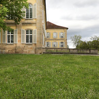Foto: Seitenansicht von Schloss Hohenheim mit gepflegtem Rasen im Vordergrund und Bäumen im Hintergrund. Das Gebäude zeigt klassische Architektur mit großen Fenstern und gelber Fassade.