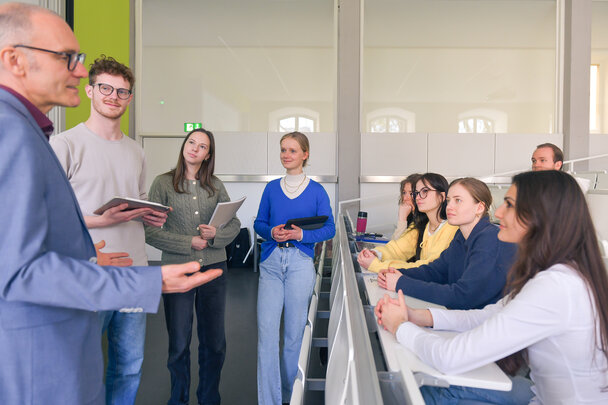 Foto: Eine Gruppe von Personen steht und sitzt in einem Hörsaal. Einige halten Tablets oder Notizbücher. Die Umgebung ist modern mit weißen Wänden und Tischen.