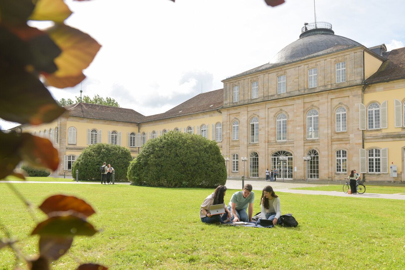 Foto: Drei Personen sitzen auf einer Wiese vor Schloss Hohenheim und arbeiten an Laptops. Im Hintergrund sind weitere Personen und das historische Gebäude sichtbar.