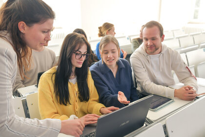 Foto: Eine Gruppe von Personen sitzt in einem Hörsaal und arbeitet gemeinsam an einem Laptop. Eine Person zeigt auf den Bildschirm, während andere aufmerksam zusehen.