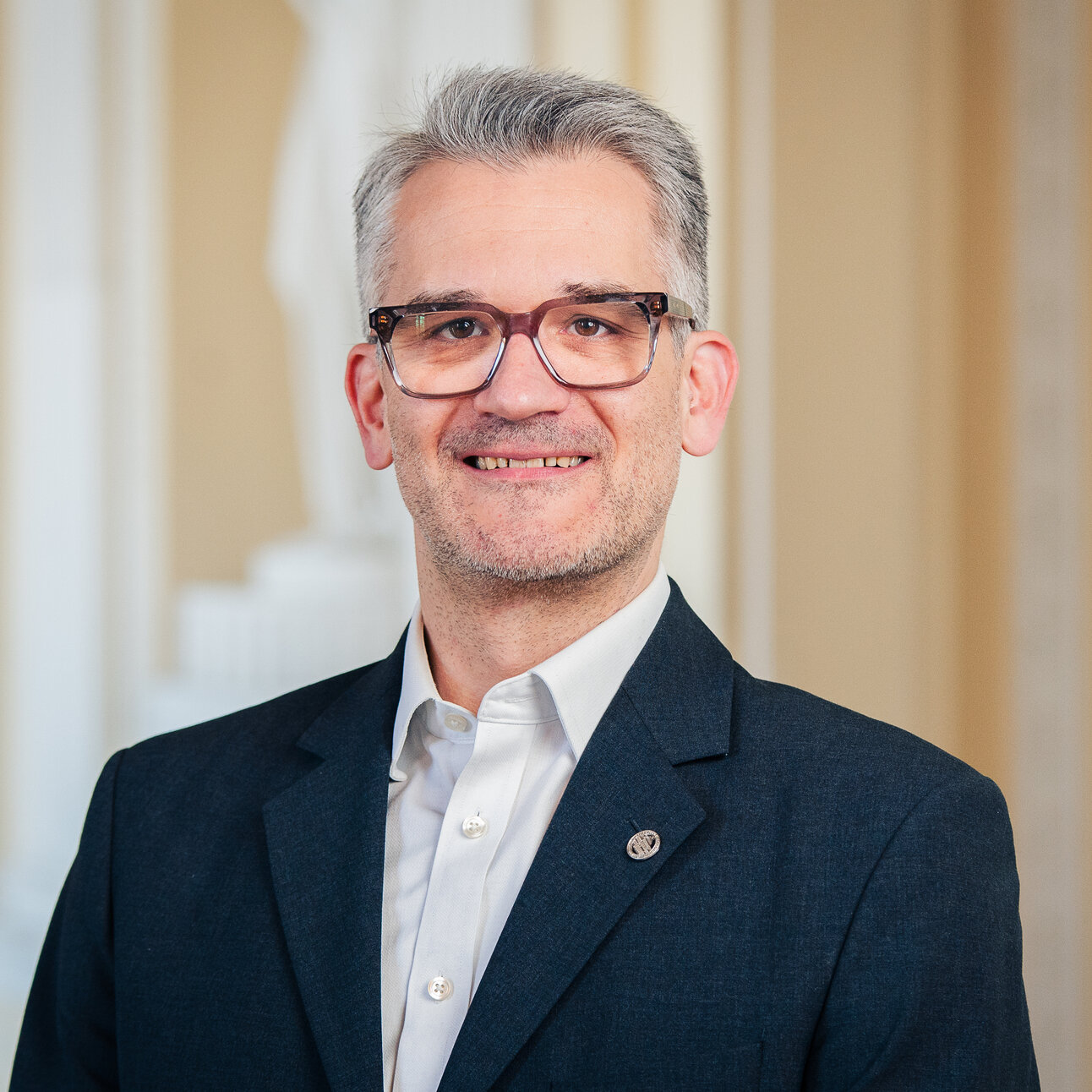 Foto: Eine Person in einem dunklen Anzug und weißem Hemd steht vor einem neutralen Hintergrund.