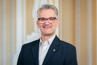 Foto: Eine Person in einem dunklen Anzug und weißem Hemd steht vor einem neutralen Hintergrund.