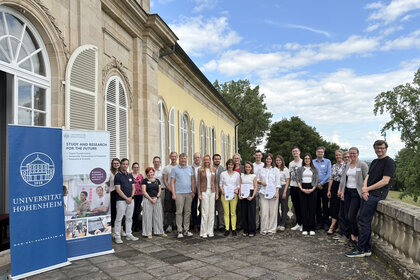 Foto: Eine Gruppe von Personen steht vor einem historischen Gebäude der Universität Hohenheim. Neben ihnen sind Banner mit der Aufschrift 'Universität Hohenheim' und 'Study and Research for the Future' zu sehen.