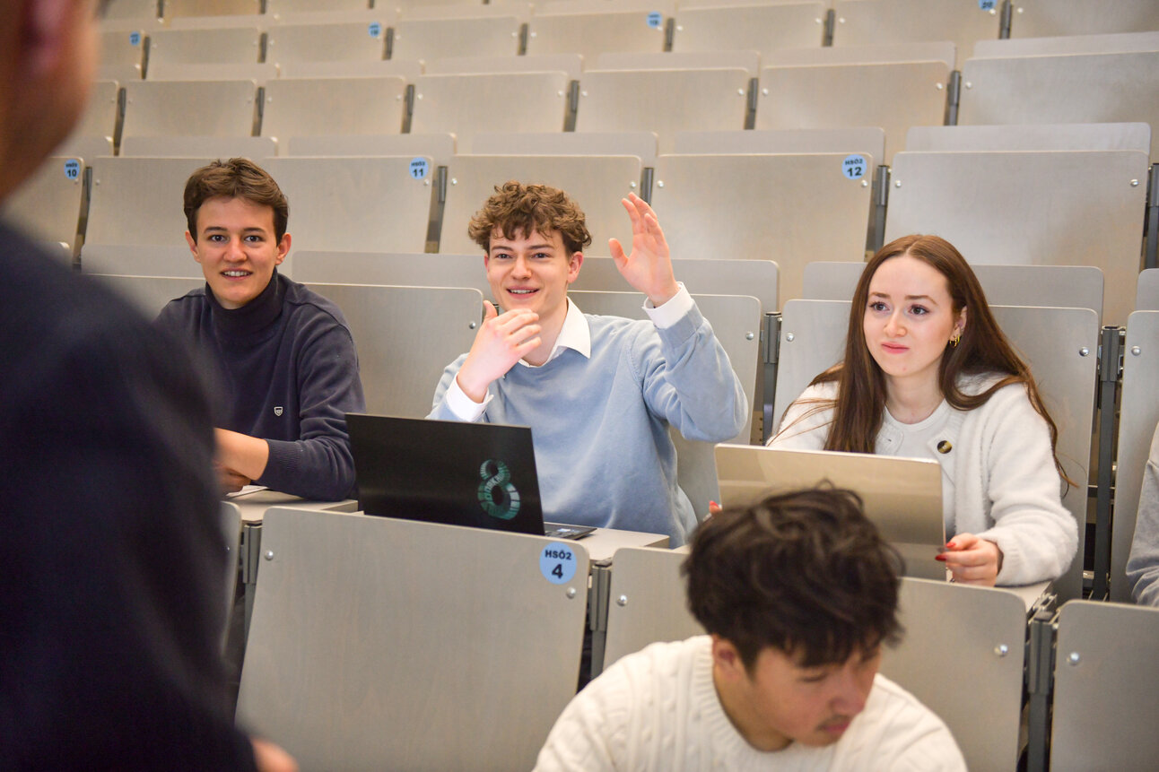 Foto: Mehrere Personen sitzen in einem Hörsaal der Universität Hohenheim. Eine Person hebt die Hand, während andere auf Laptops arbeiten. Die Sitzreihen sind nummeriert.