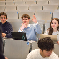Foto: Mehrere Personen sitzen in einem Hörsaal der Universität Hohenheim. Eine Person hebt die Hand, während andere auf Laptops arbeiten. Die Sitzreihen sind nummeriert.