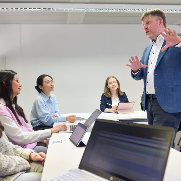 Foto: Eine Person in einem blauen Blazer spricht zu mehreren Personen, die an einem Tisch mit Laptops sitzen. Ein Flipchart mit Diagrammen ist im Hintergrund sichtbar.