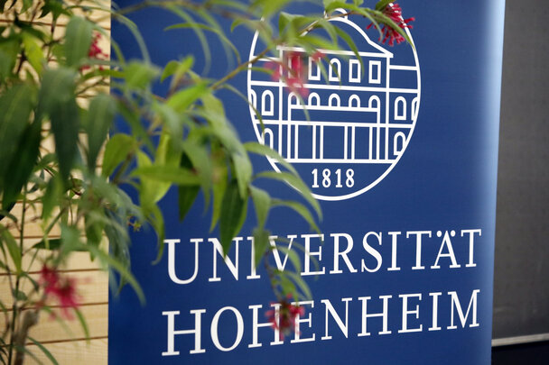 Foto: Ein blaues Banner mit dem Logo der Universität Hohenheim und der Jahreszahl 1818, teilweise verdeckt von Pflanzen im Vordergrund.