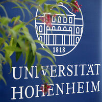 Foto: Ein blaues Banner mit dem Logo der Universität Hohenheim und der Jahreszahl 1818, teilweise verdeckt von Pflanzen im Vordergrund.