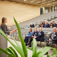 Foto: Eine Person hält einen Vortrag in einem Hörsaal vor einem Publikum. Die Zuhörer sitzen in Reihen auf ansteigenden Sitzplätzen. Pflanzen sind im Vordergrund sichtbar.