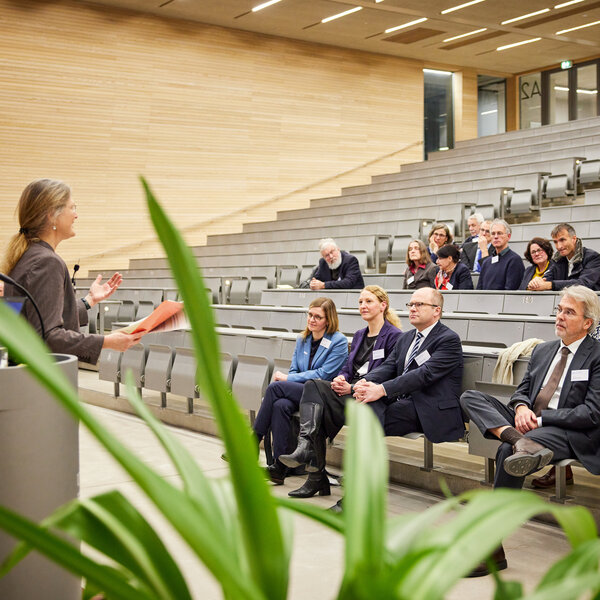 Foto: Eine Person hält einen Vortrag in einem Hörsaal vor einem Publikum. Die Zuhörer sitzen in Reihen auf ansteigenden Sitzplätzen. Pflanzen sind im Vordergrund sichtbar.