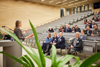 Foto: Eine Person hält einen Vortrag in einem Hörsaal vor einem Publikum. Die Zuhörer sitzen in Reihen auf ansteigenden Sitzplätzen. Pflanzen sind im Vordergrund sichtbar.
