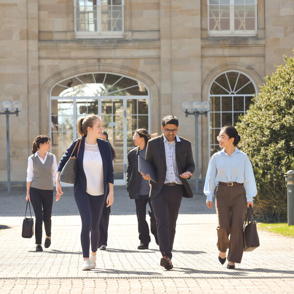 Foto: Eine Gruppe von Personen geht auf einem gepflasterten Weg vor dem Schloss Hohenheim. Das Gebäude im Hintergrund hat große Fenster und klassische Architektur.