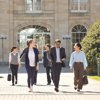 Foto: Eine Gruppe von Personen geht auf einem gepflasterten Weg vor dem Schloss Hohenheim. Das Gebäude im Hintergrund hat große Fenster und klassische Architektur.