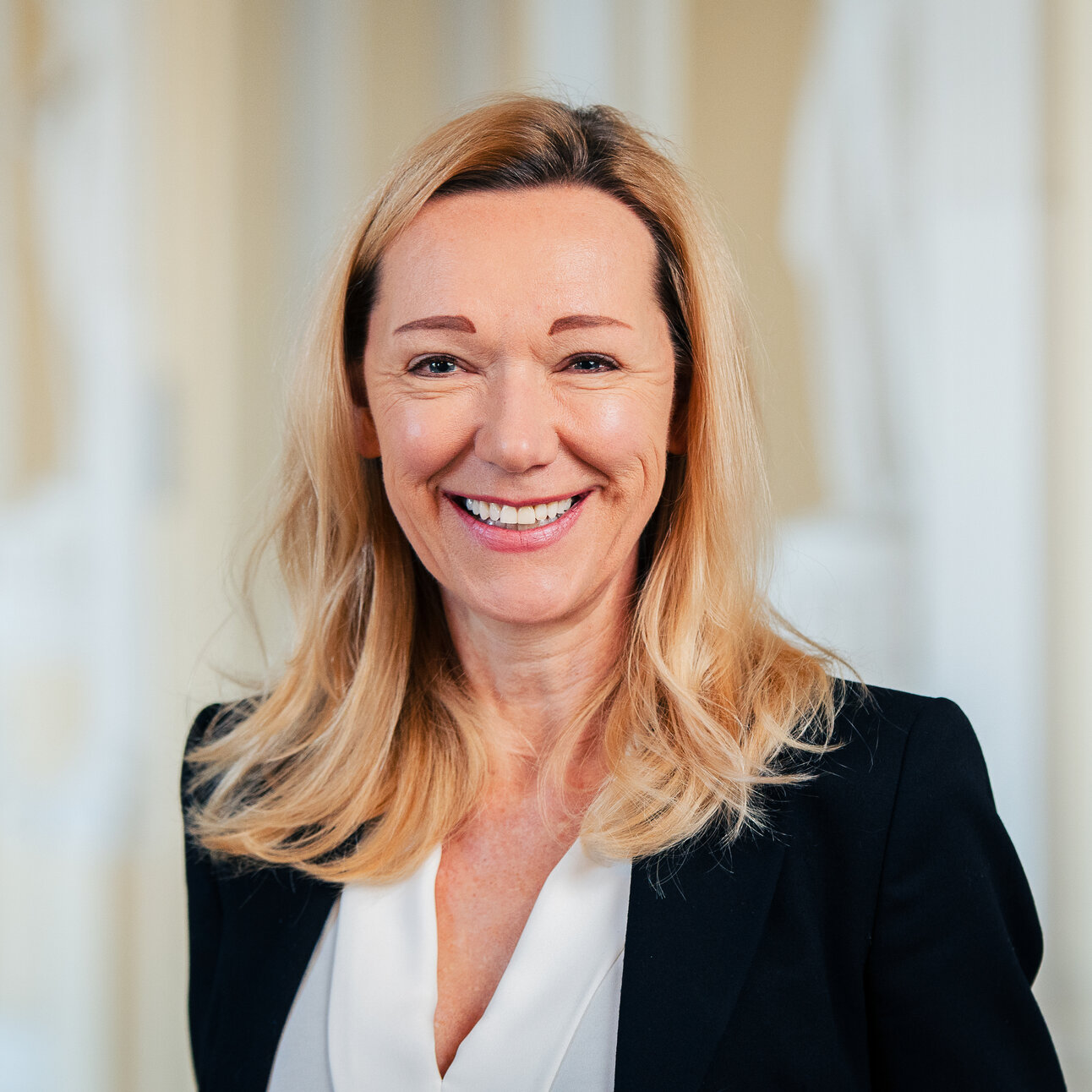 Foto: Eine Person in einem schwarzen Blazer und weißer Bluse steht vor einem neutralen Hintergrund in einem Innenraum.