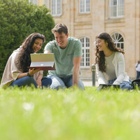 Foto: Drei Personen sitzen auf einer Wiese vor einem großen Gebäude und schauen auf einen Laptop. Im Hintergrund sind weitere Personen und das Gebäude sichtbar.