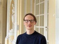 Foto: Eine Person steht in einem historischen Gebäude mit dekorativen Säulen und einer Statue im Hintergrund. Die Person trägt einen dunklen Pullover und steht vor einer weißen Tür.