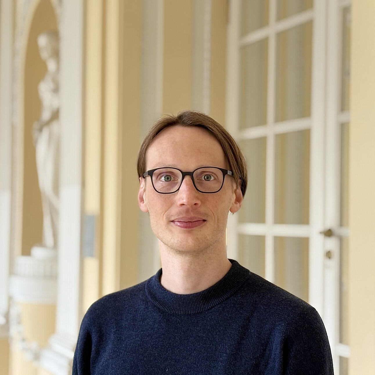 Foto: Eine Person steht in einem historischen Gebäude mit dekorativen Säulen und einer Statue im Hintergrund. Die Person trägt einen dunklen Pullover und steht vor einer weißen Tür.