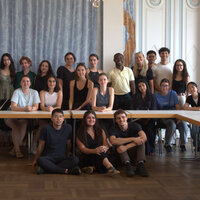 Foto: Eine Gruppe von Personen sitzt und steht in einem Raum mit blauen Vorhängen und dekorativen Wänden. Die Personen sind um einen Tisch gruppiert, der in U-Form angeordnet ist.