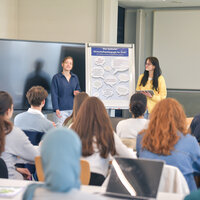 Foto: Zwei Personen präsentieren vor einer Gruppe von Studierenden. Ein Flipchart zeigt den Text 'Was bedeutet Wirtschaftspädagogik für Dich?'.