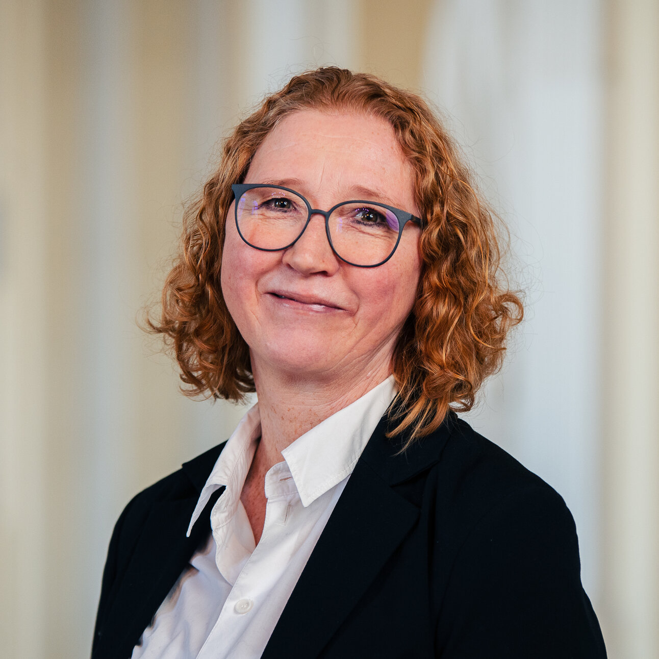 Foto: Eine Person in einem dunklen Blazer und weißem Hemd steht vor einem neutralen Hintergrund.