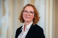 Foto: Eine Person in einem dunklen Blazer und weißem Hemd steht vor einem neutralen Hintergrund.