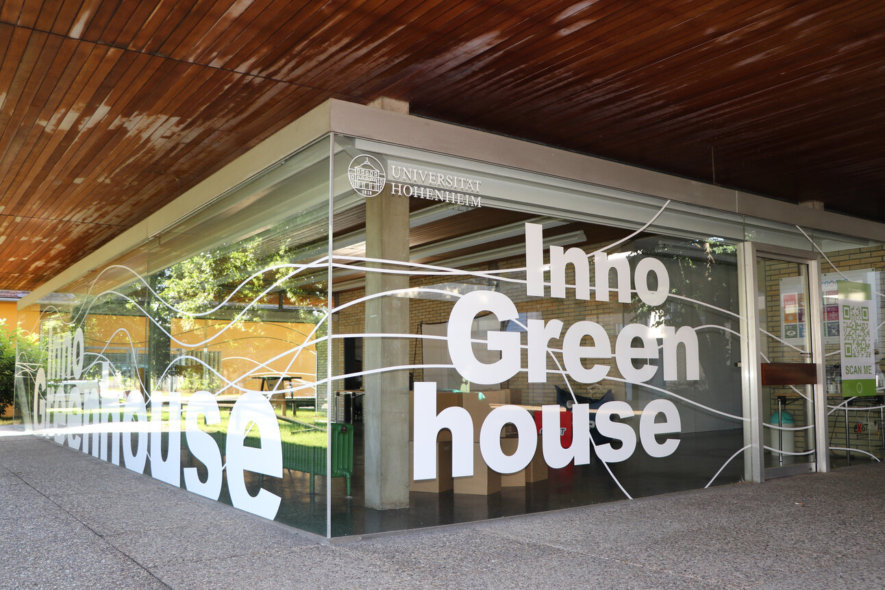 Foto: Das Inno Greenhouse der Universität Hohenheim mit großen Glaswänden und der Aufschrift 'Inno Greenhouse'. Im Inneren sind Pflanzen und Möbel sichtbar.