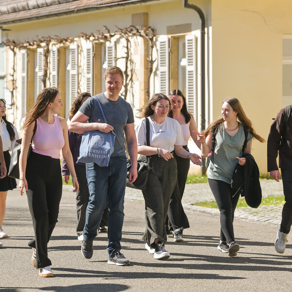 Foto: Eine Gruppe von Personen geht auf einem Gehweg vor einem Gebäude auf dem Campus der Universität Hohenheim. Eine Person trägt eine Tasche mit dem Logo der Universität.