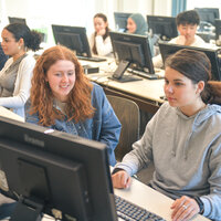 Foto: Mehrere Personen sitzen in einem Computerraum an Schreibtischen mit Monitoren. Sie arbeiten an Computern in einer Lernumgebung.