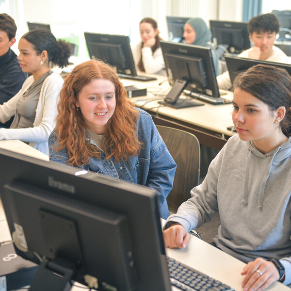 Foto: Mehrere Personen sitzen in einem Computerraum an Schreibtischen mit Monitoren. Sie arbeiten an Computern in einer Lernumgebung.