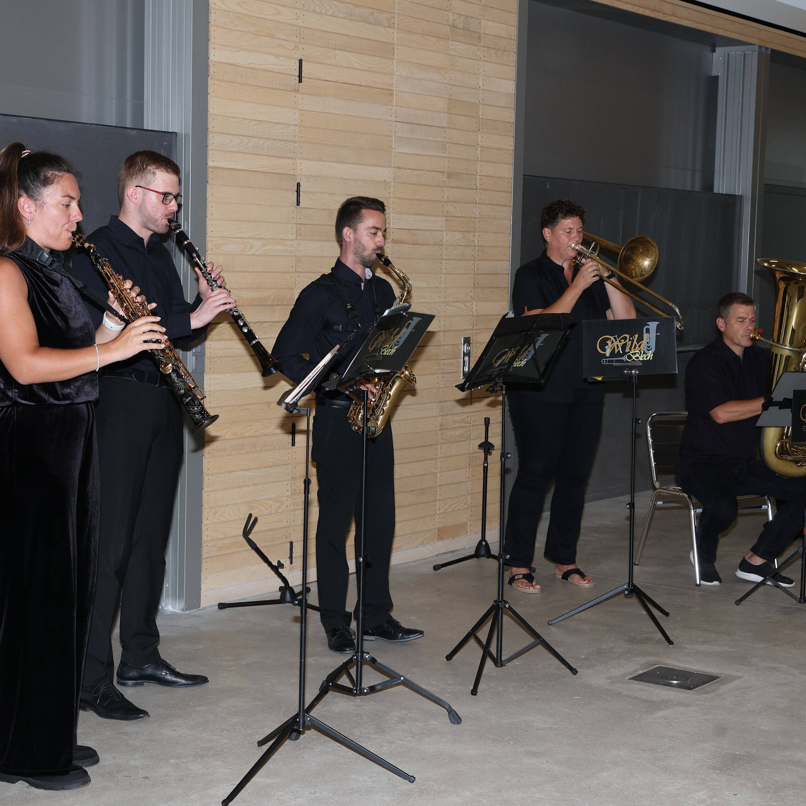 Foto: Eine Gruppe von Personen spielt Musikinstrumente in einem Innenraum. Sie verwenden Klarinetten, Saxophon, Posaune und Tuba. Notenständer mit dem Logo 'Wild Salon' sind vor ihnen aufgestellt.