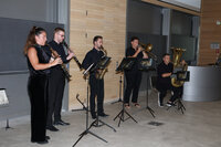 Foto: Eine Gruppe von Personen spielt Musikinstrumente in einem Innenraum. Sie verwenden Klarinetten, Saxophon, Posaune und Tuba. Notenständer mit dem Logo 'Wild Salon' sind vor ihnen aufgestellt.