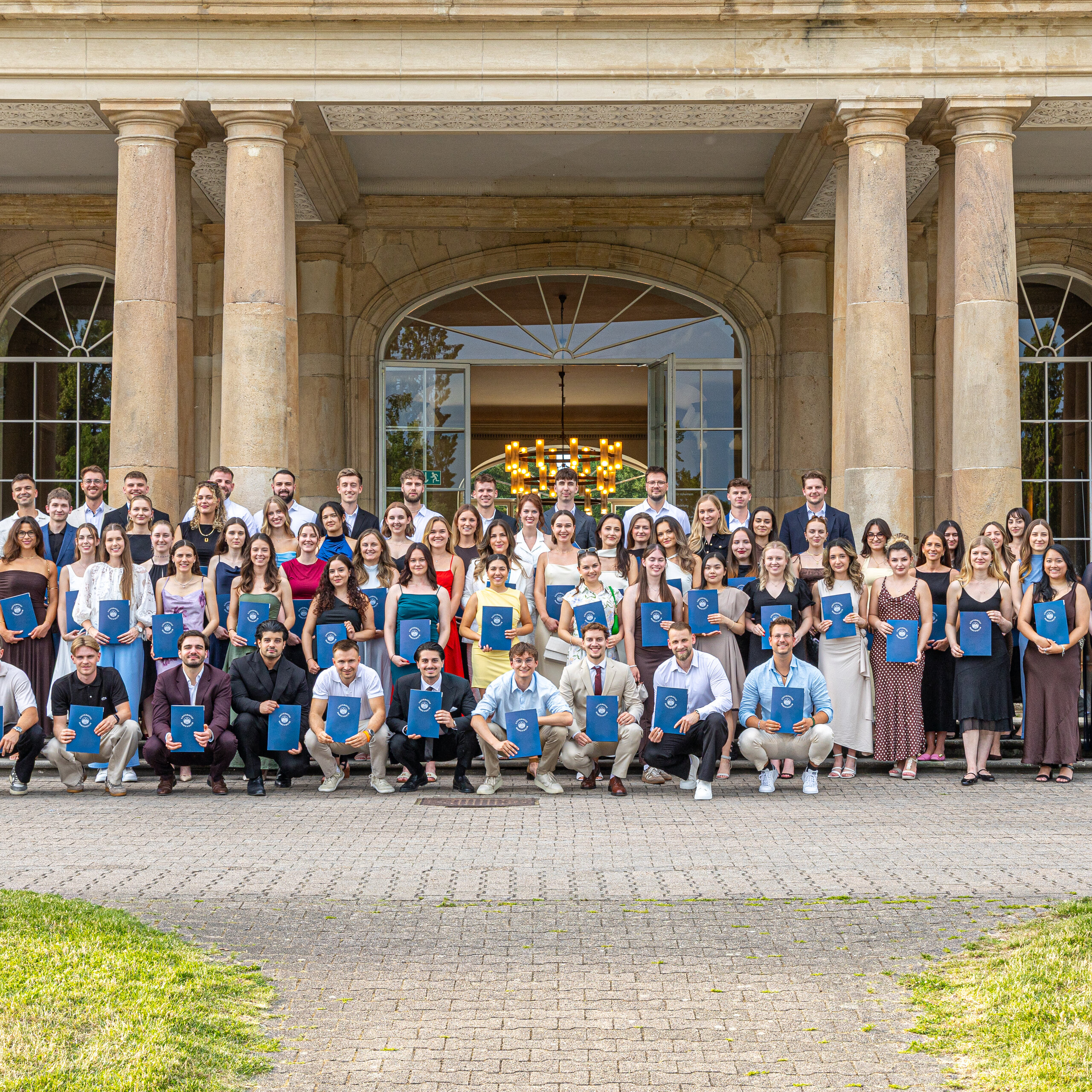 Foto: Eine Gruppe von Personen steht vor Schloss Hohenheim. Sie halten Zertifikate und posieren für ein Foto. Das Gebäude im Hintergrund hat Säulen und große Fenster.