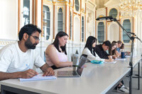 Foto: Mehrere Personen sitzen an einem Tisch in einem dekorativen Raum und machen Notizen. Laptops und Schreibmaterialien sind auf dem Tisch verteilt.