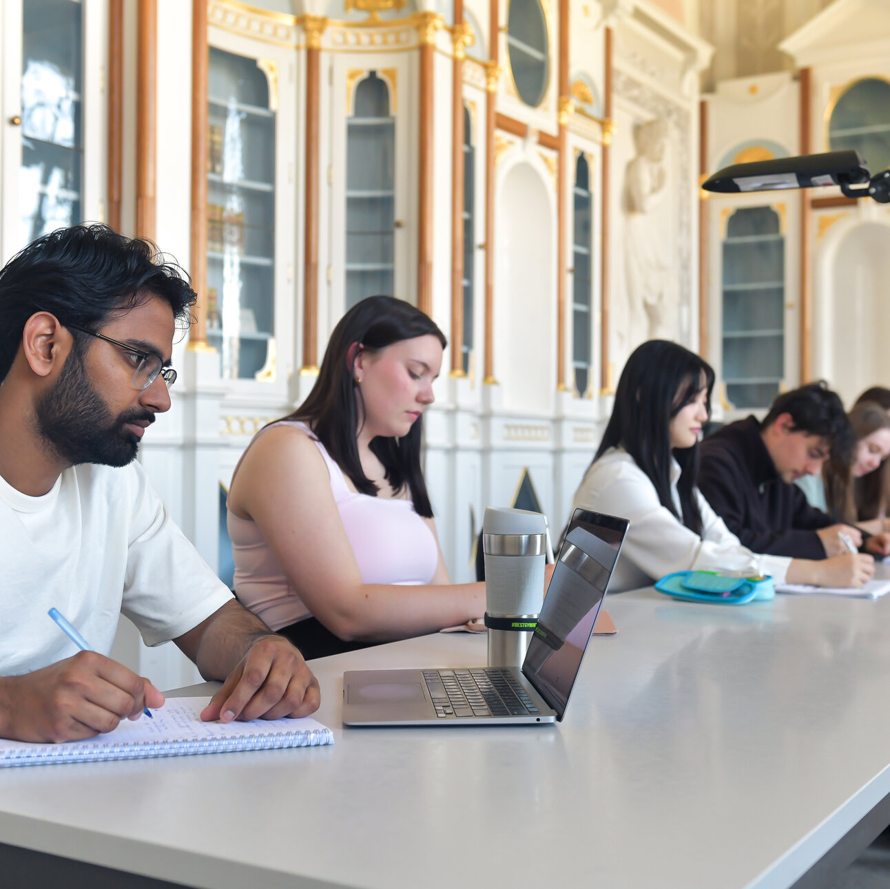 Foto: Mehrere Personen sitzen an einem Tisch in einem dekorativen Raum und machen Notizen. Laptops und Schreibmaterialien sind auf dem Tisch verteilt.