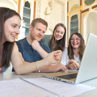 Foto: Eine Gruppe von Personen sitzt an einem Tisch und arbeitet gemeinsam an einem Laptop. Im Hintergrund sind dekorative Elemente und Regale sichtbar.